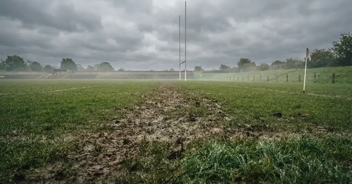 Boisko do rugby ze słupami bramki w oddali pod pochmurnym niebem, mokra trawa i ślady butów.